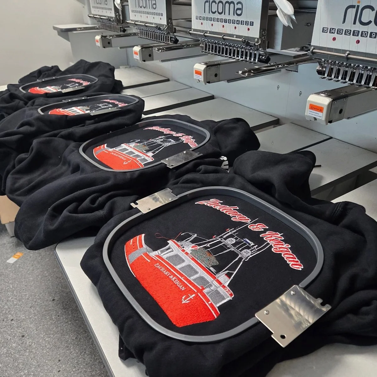 Black garments held in embroidery hoops beneath machine needles for logo stitching